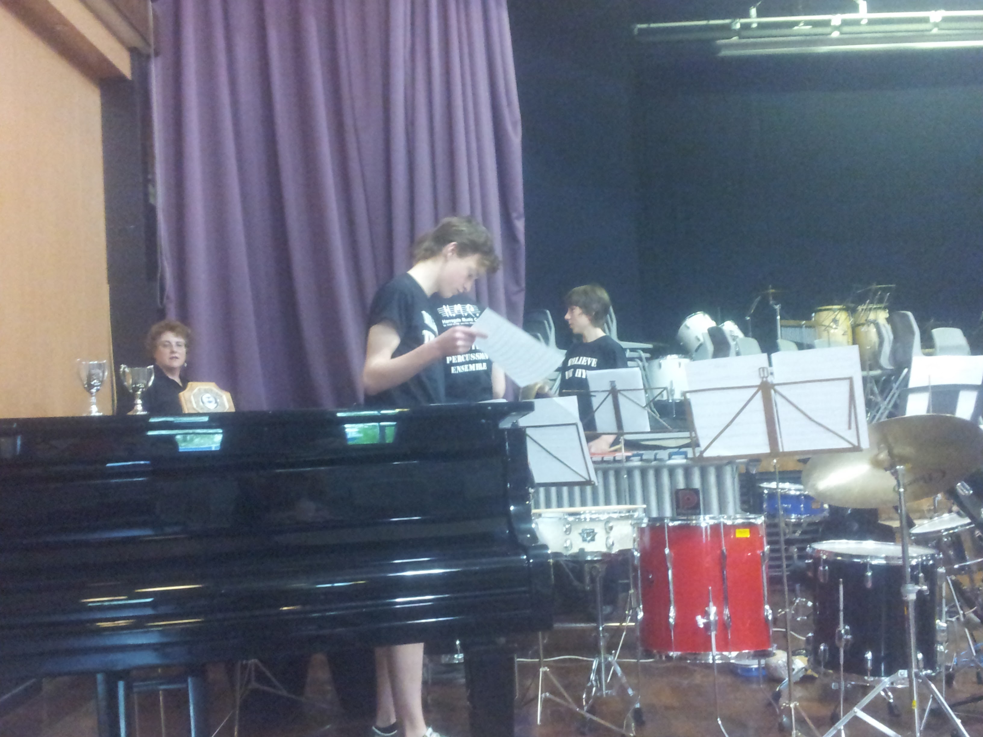 A stage with a piano, a drum kit and percussionists in black T-shirts standing by music stands