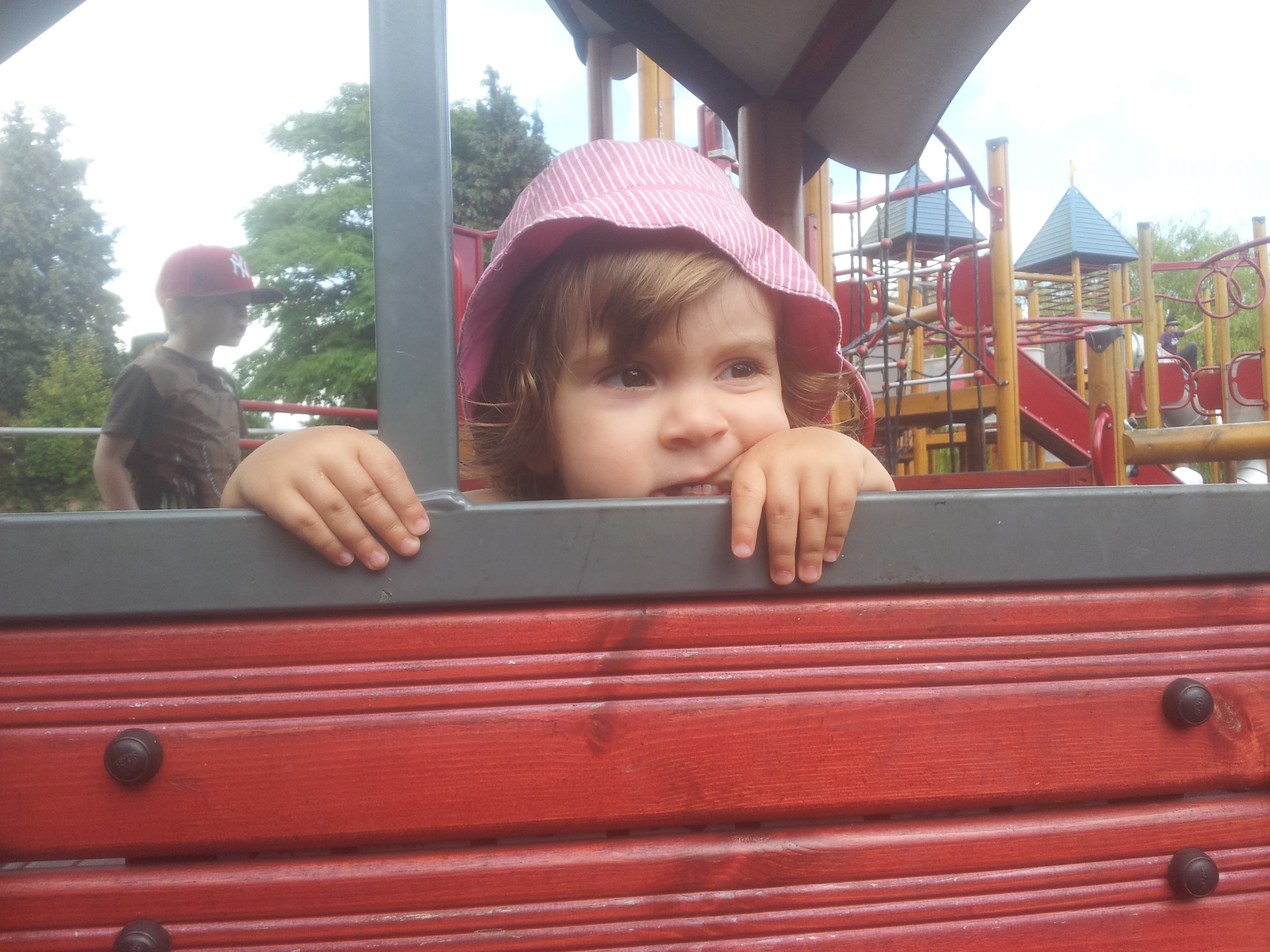 A toddler in a pink sun hat looks over a red wooden panel at a playground