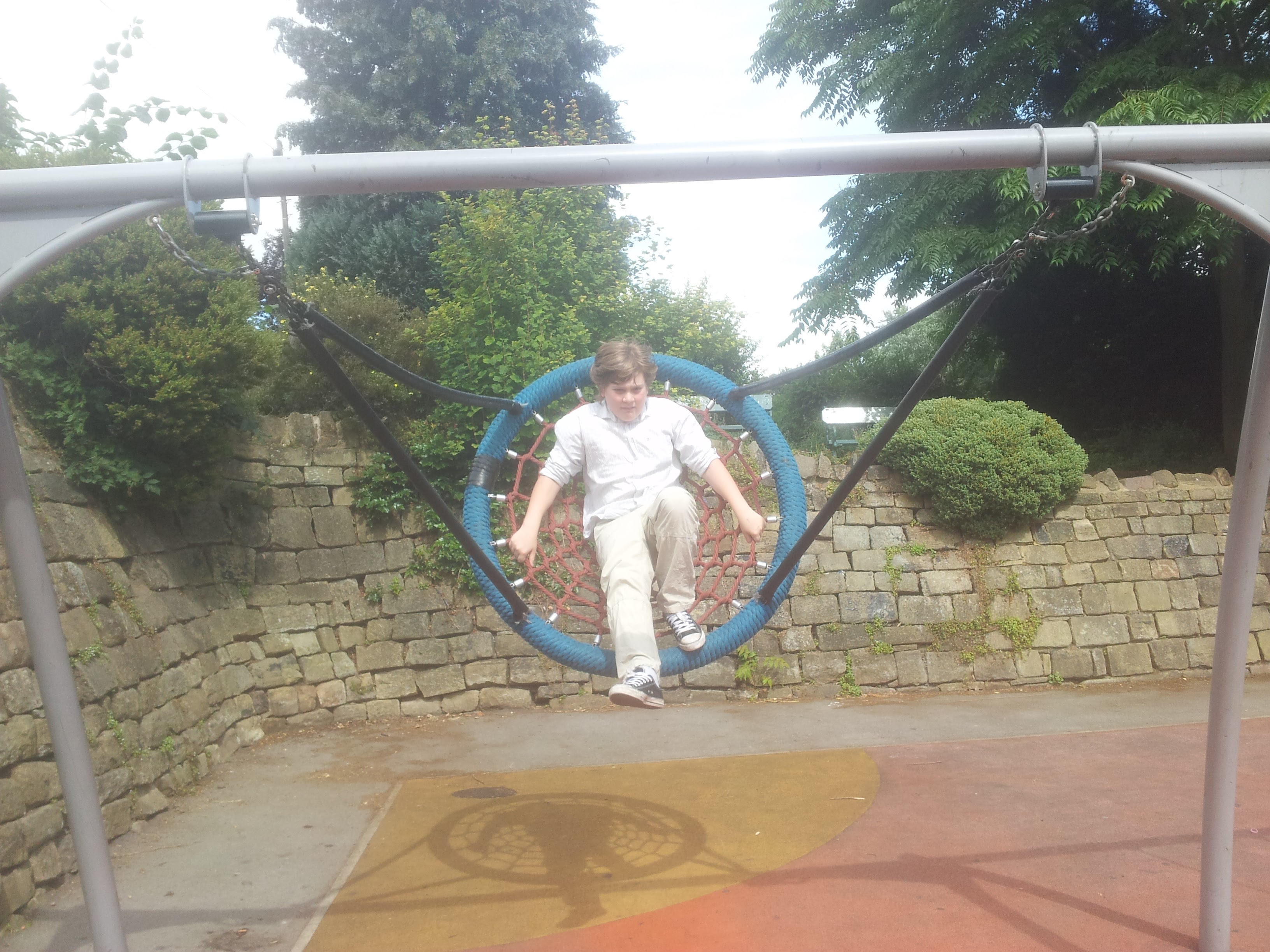A child stands inside a large circular rope swing in a playground beside a stone wall