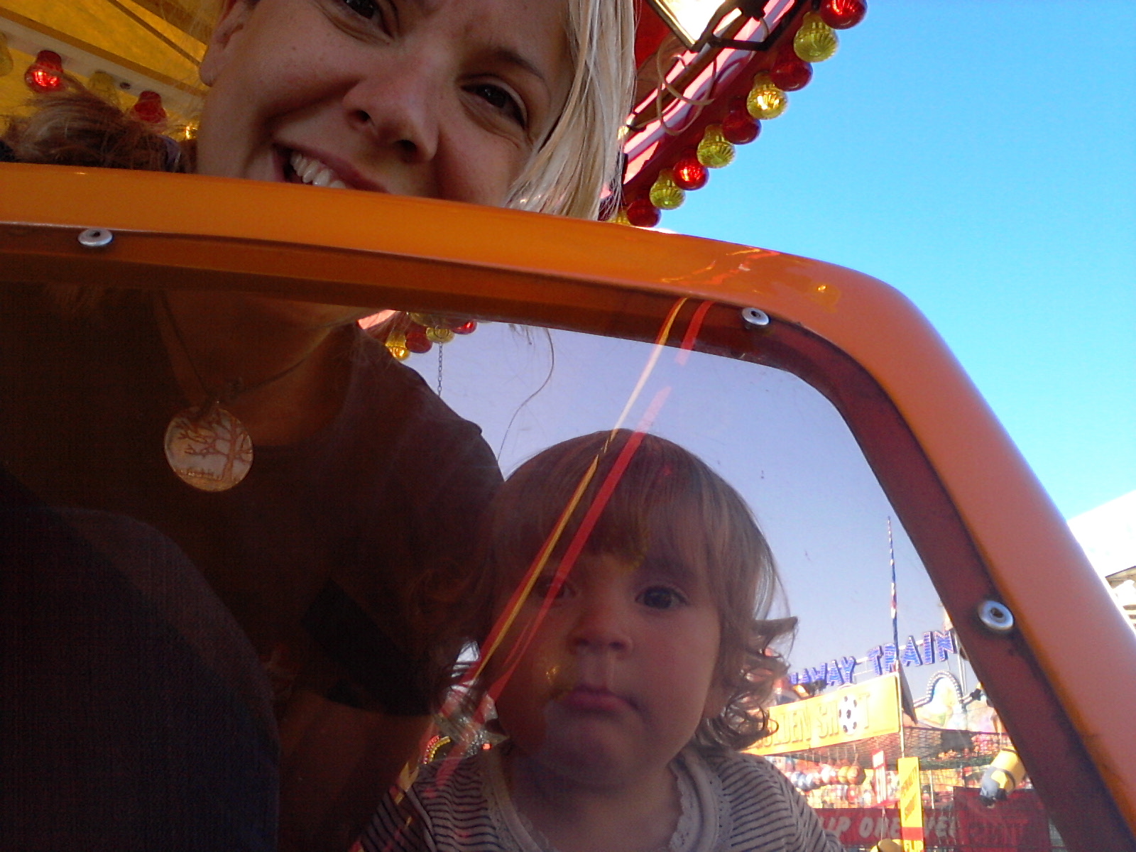 A smiling adult leans in over a toddler inside an orange fairground car