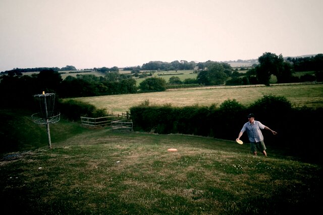 Person throws a disc on a grassy hillside with a disc golf basket beside them