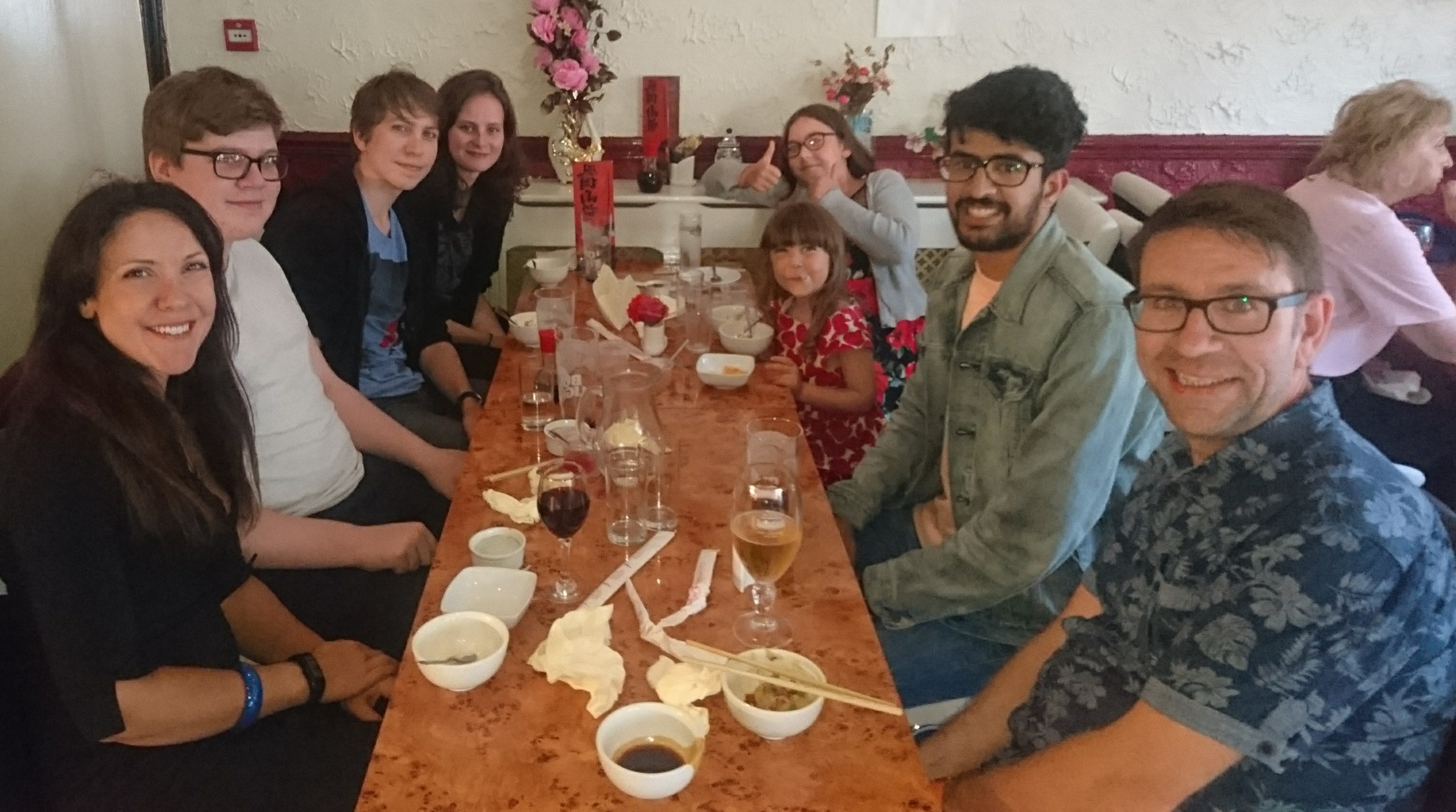 Eight people seated along a wooden table in a restaurant, smiling at the camera, one child giving a thumbs up