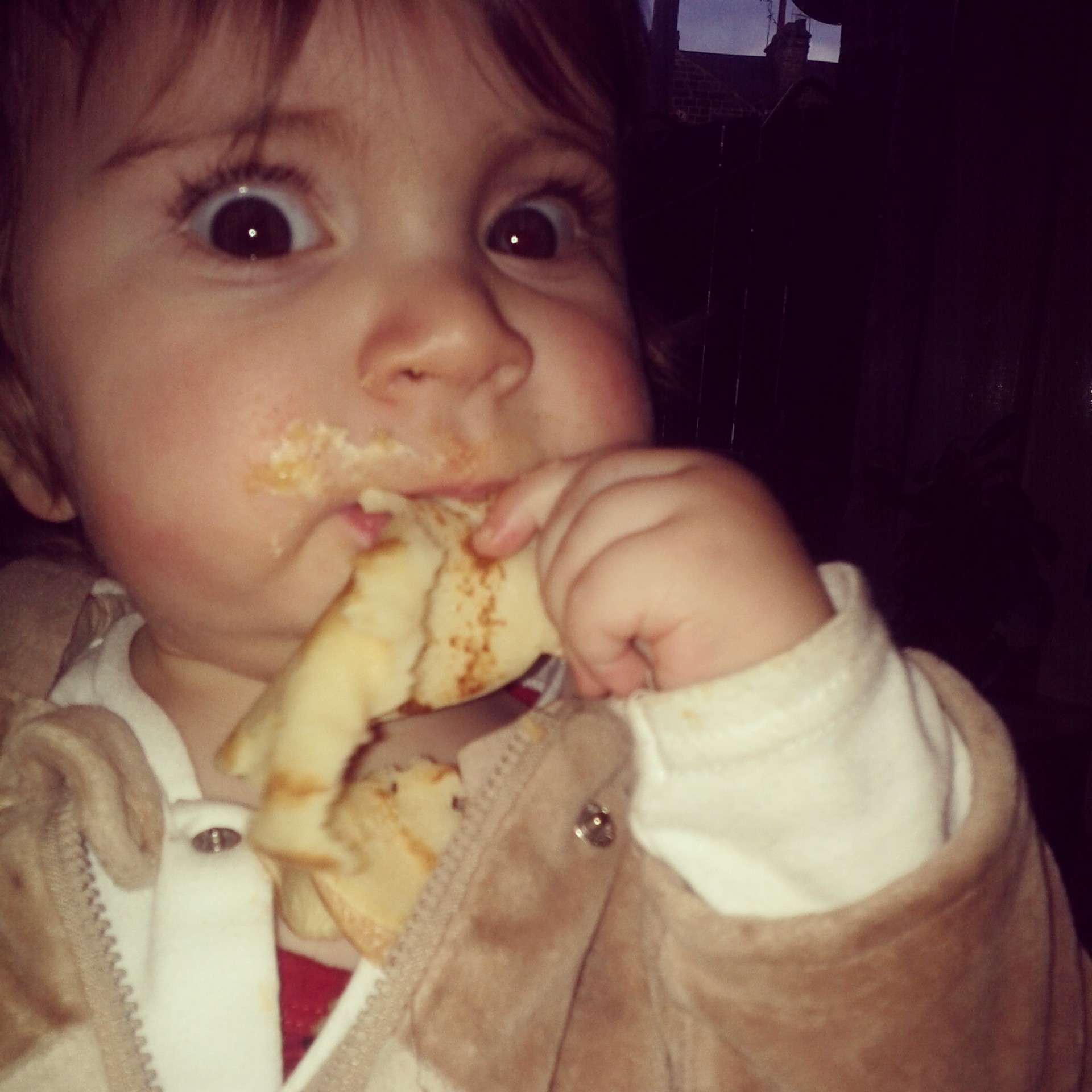 A baby with wide eyes eats a pancake, with food around the mouth