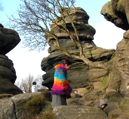A person in a rainbow knitted coat stands among weathered rocks and a bare tree at Brimham Rocks