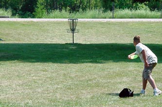Person throws a frisbee towards a disc golf basket on a lawn, with a black bag on the grass