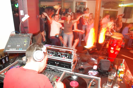 DJ in a red T-shirt and headphones at the decks with a crowd dancing behind and flames