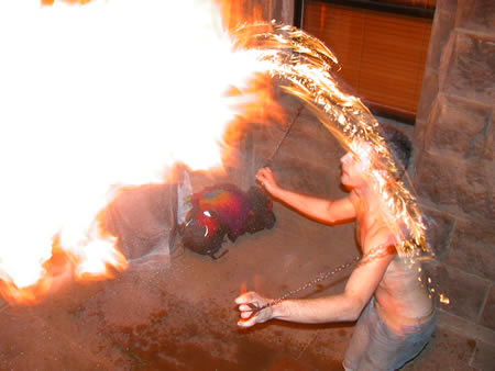 Shirtless person spinning a flaming rope or chain beside a stone building, with flames to the left