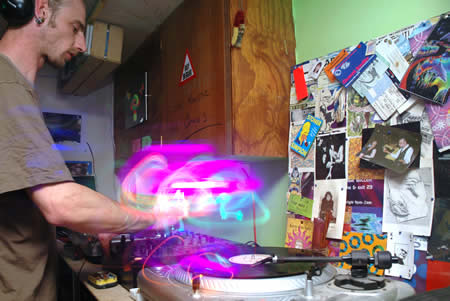 DJ in a T-shirt mixing on a turntable with a pink light blur, posters on the wall behind