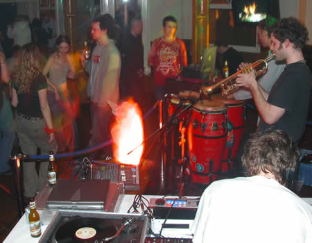 A trumpeter and conga player perform by DJ decks as people stand around in a bar
