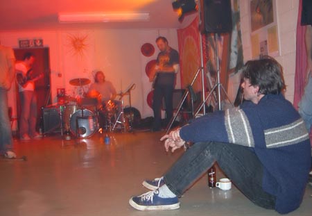 A person in a blue jumper sits on the floor while a band plays in a red-lit room behind