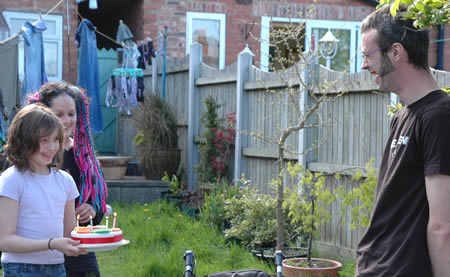 Child holding a birthday cake with candles in a garden, with two adults nearby
