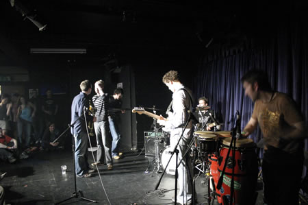 Band playing on a dark stage with a guitarist, drummer and a conga player, audience sitting at the left
