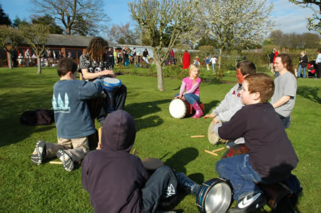 People sit on grass playing hand drums in a park, with a child in pink in the background