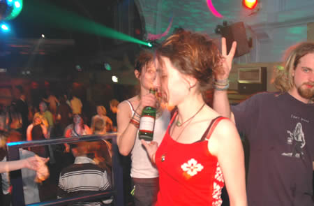A woman in a red top holds a bottle while a bearded man makes a peace sign in a club