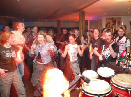 People dance in a crowded bar beside congas, with a burst of flame in the foreground