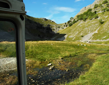 Grassy limestone valley with a stream, seen from inside a vehicle window