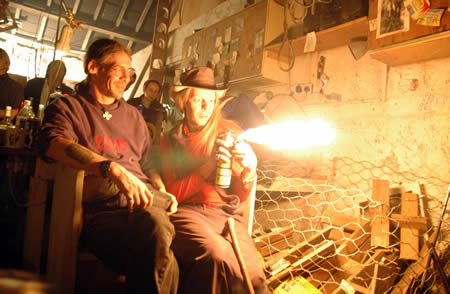Two people sit in a cluttered wooden shed while a flame shoots from a spray can