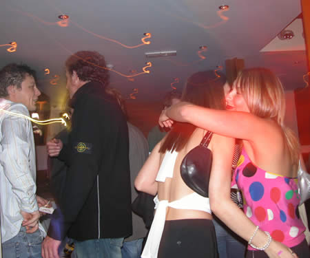 Two women hugging in a crowded bar, with a man in a dark jacket beside them