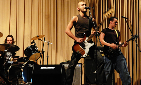 Two bass players and a drummer perform on a stage with gold curtains