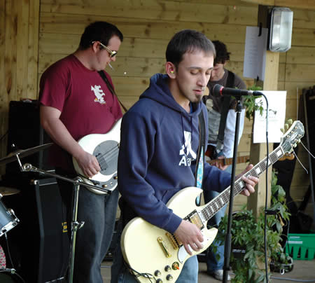 Two guitarists play in a wooden shelter, one in a maroon T-shirt and one in a navy hoodie