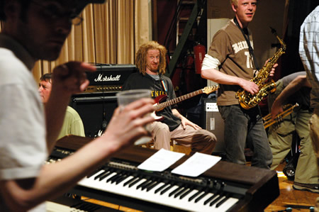 Band in a hall with a keyboardist in the foreground, a saxophonist and a guitarist behind