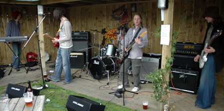 Band plays in a wooden open-fronted shelter with amps, a drum kit and a keyboard