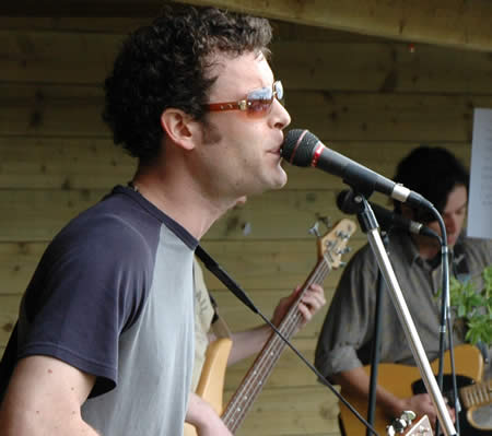 Singer with curly hair and sunglasses at a microphone in a wooden shelter, bassist behind