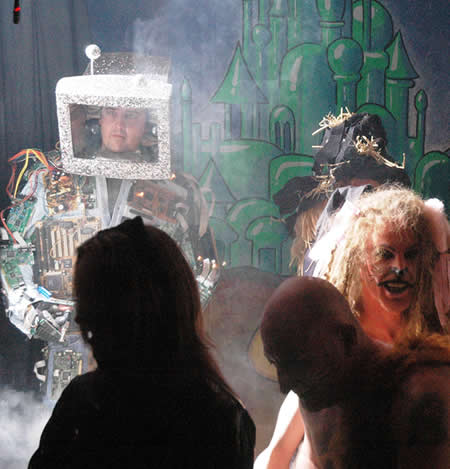 Person with a computer-monitor head costume beside a scarecrow and a lion in make-up, in front of a green city backdrop