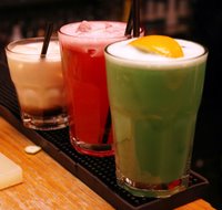 Three tall cocktails on a bar, one cream, one red and one green with an orange slice
