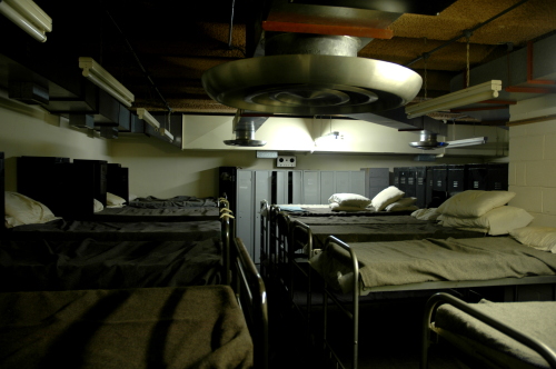 Rows of metal-framed beds with grey blankets in a low underground dormitory