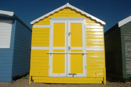 Yellow beach hut with white double doors between blue and green huts