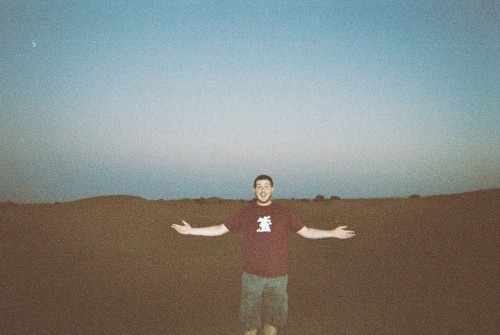 A person in a maroon T-shirt stands with arms outstretched in a barren landscape at dusk