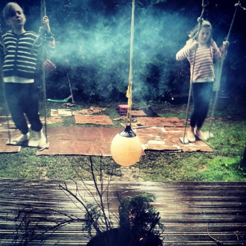 Two girls stand on rope swings in a smoky garden, a round white lamp hanging between them
