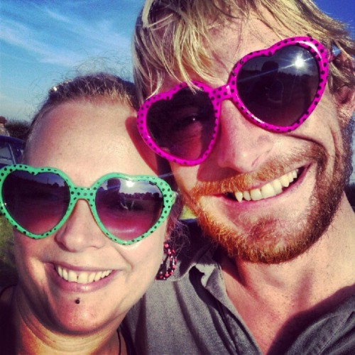 Two smiling people in heart-shaped sunglasses, one pink and one green, taking a selfie