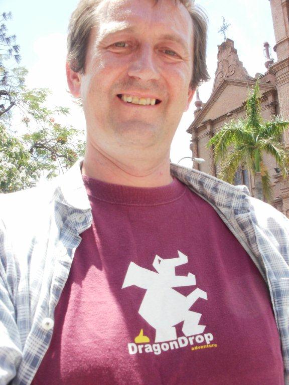 Smiling man in a maroon DragonDrop T-shirt and checked shirt, with a church behind