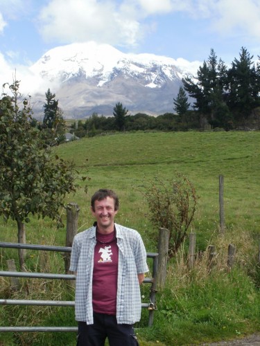 A man in a maroon T-shirt and checked shirt stands by a fence, with a snow-capped mountain behind