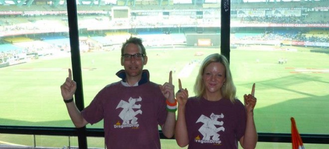 Two people in matching maroon DragonDrop t-shirts point upwards in front of a cricket stadium