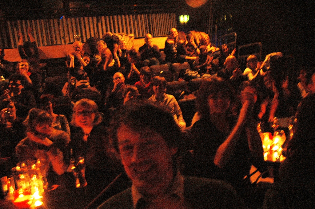 Dimly lit audience seated in a theatre with candles on tables, and a smiling man in the foreground