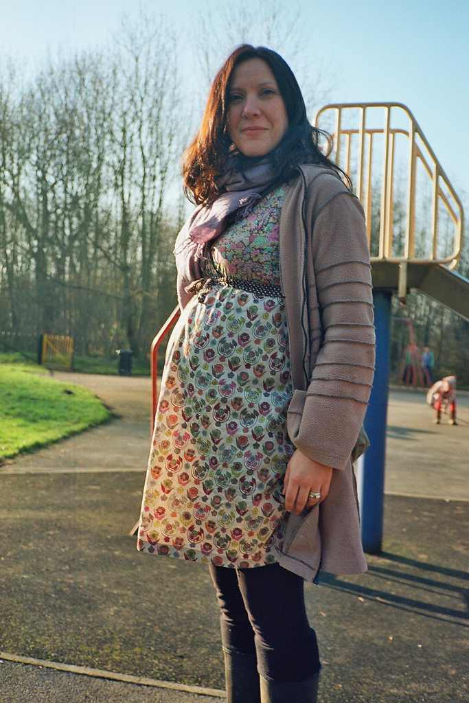 Pregnant person in a floral dress and cardigan stands in a playground