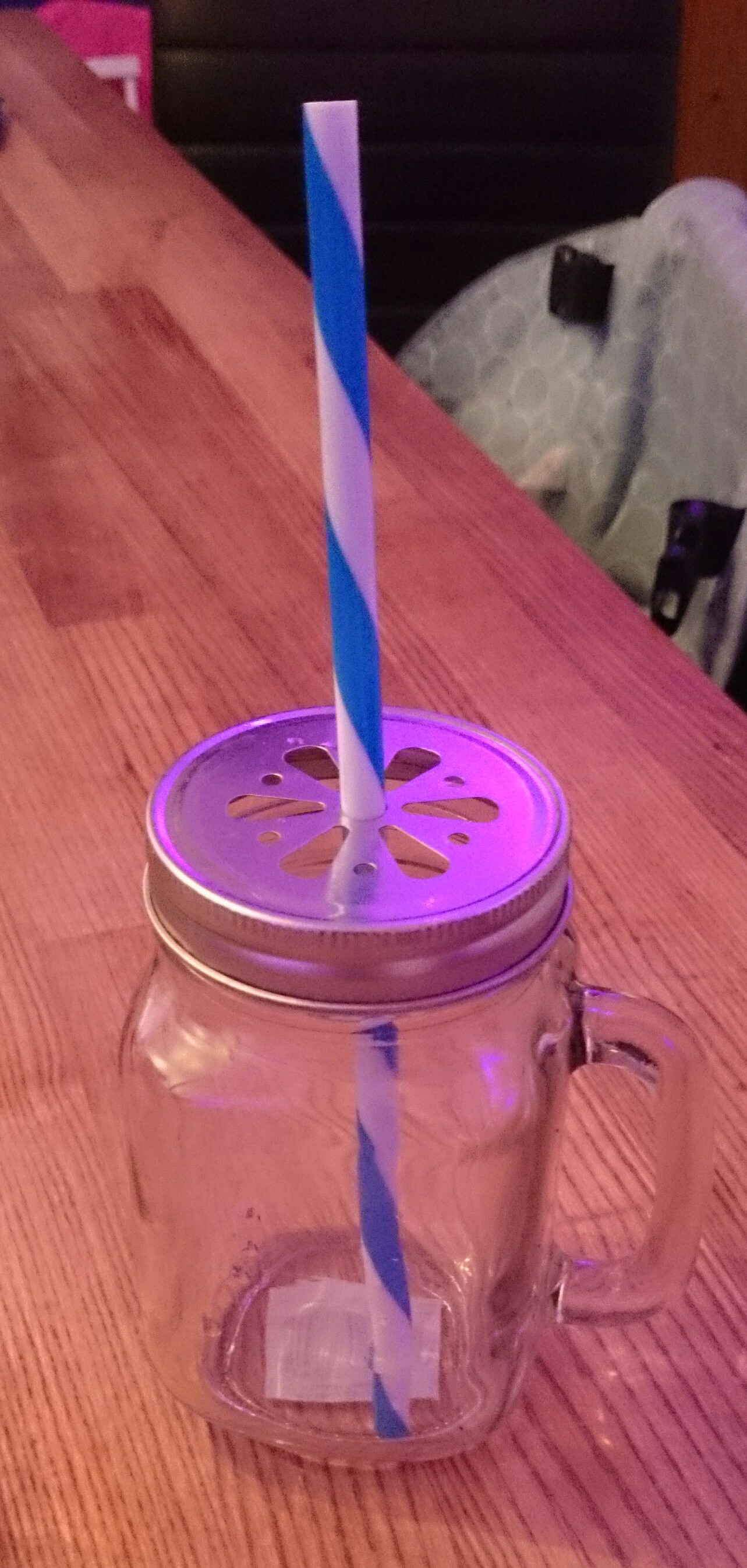 A glass jar with a handle, a metal lid and a blue and white striped straw on a wooden bar