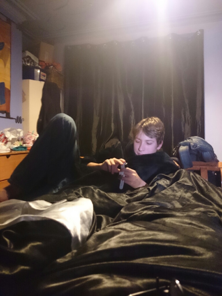 A teenager sits up in bed in a dim room, looking at a phone under a black duvet