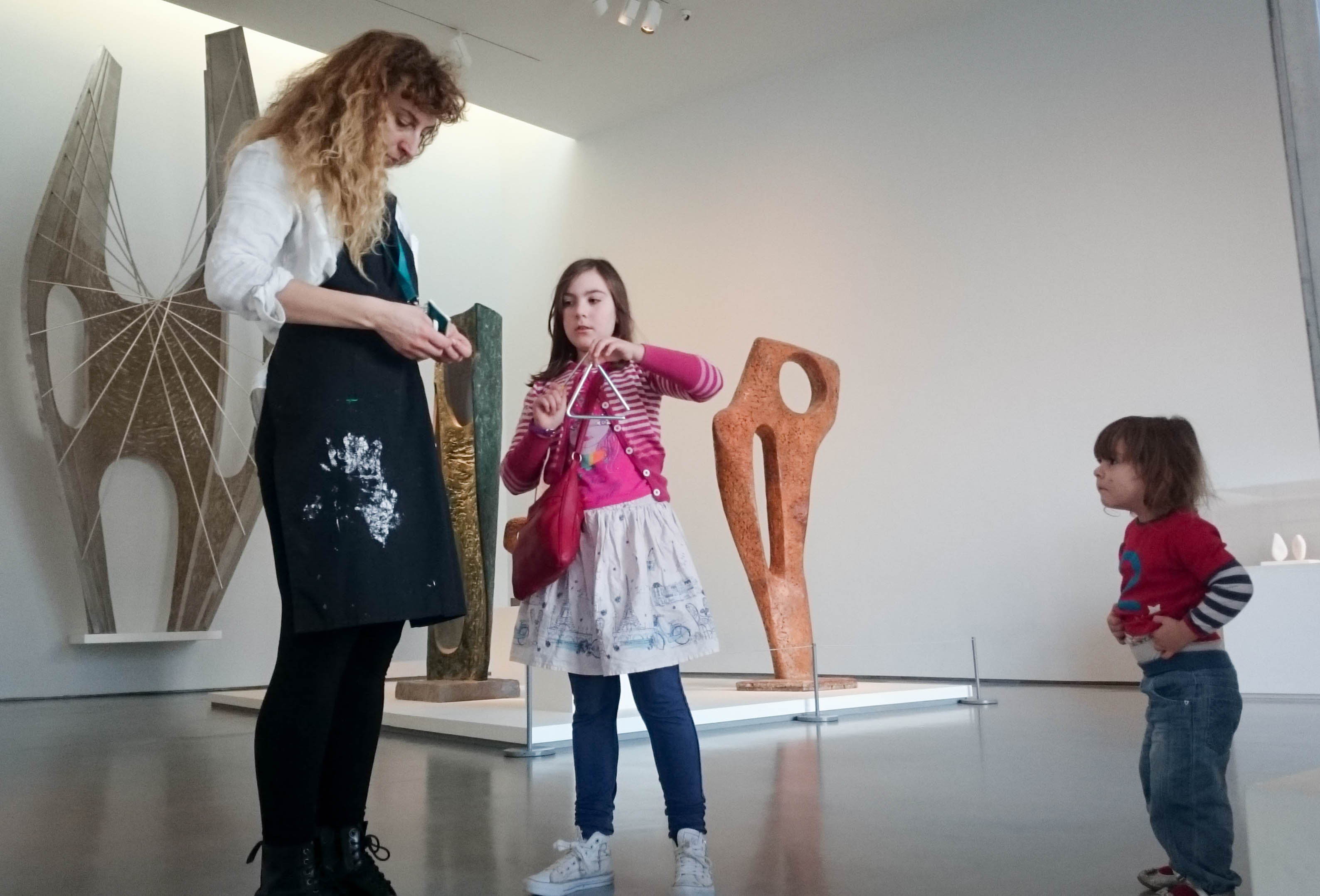 A woman in an apron and a child stand in a gallery near tall sculptures while a toddler looks on