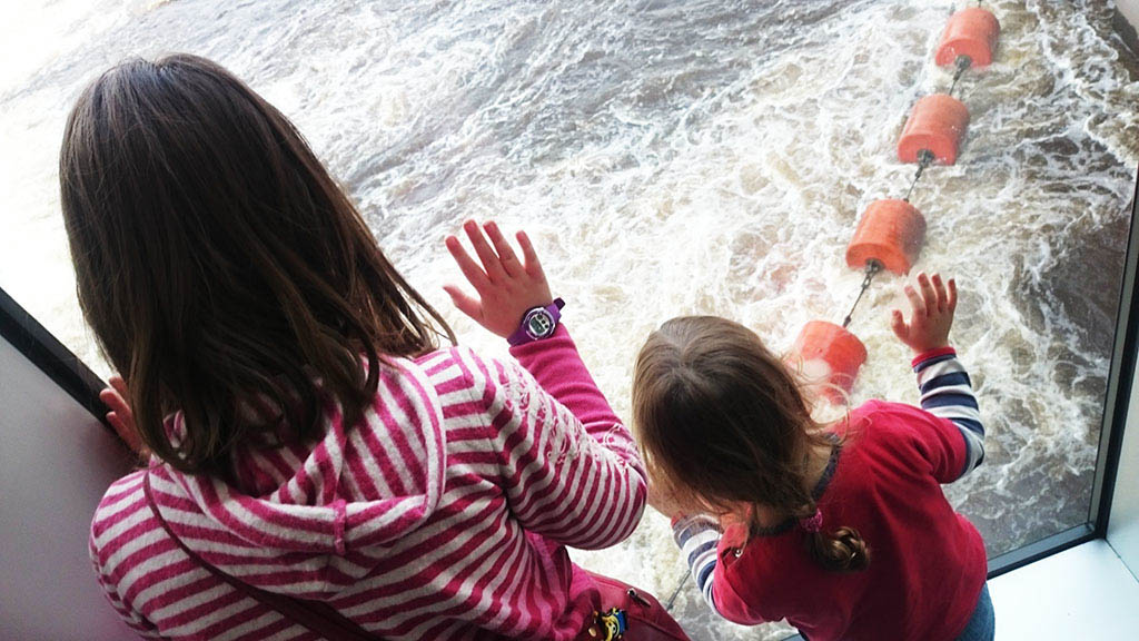 Two children in a striped top and a red top look out of a window at churning water with orange floats