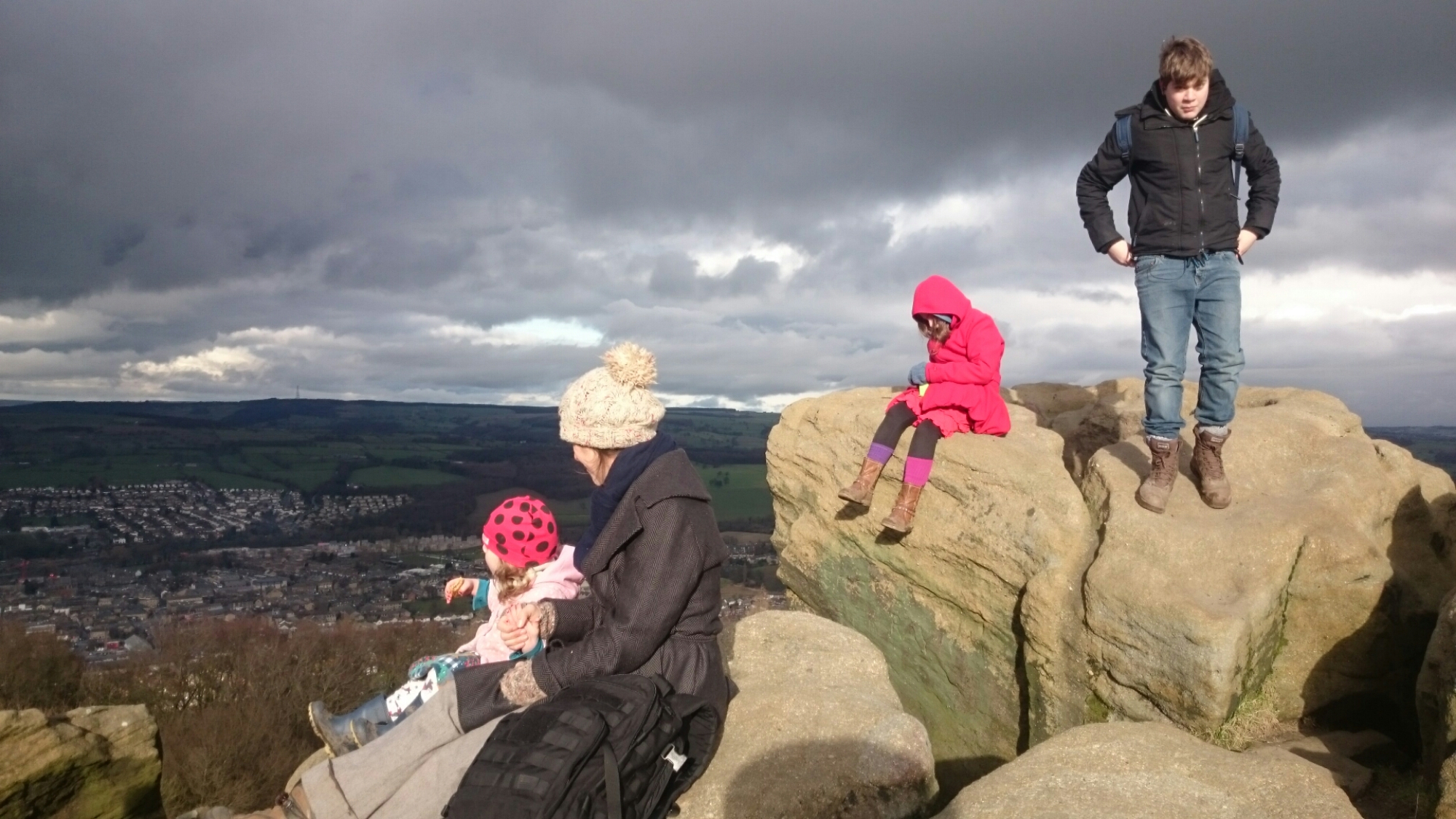A family sits and stands on gritstone rocks on a hilltop under a cloudy sky, with a town below