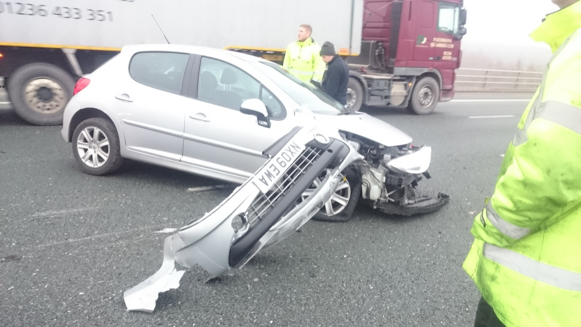 A silver car with a wrecked front and a detached bumper on a road, with a lorry and two people in hi-vis behind
