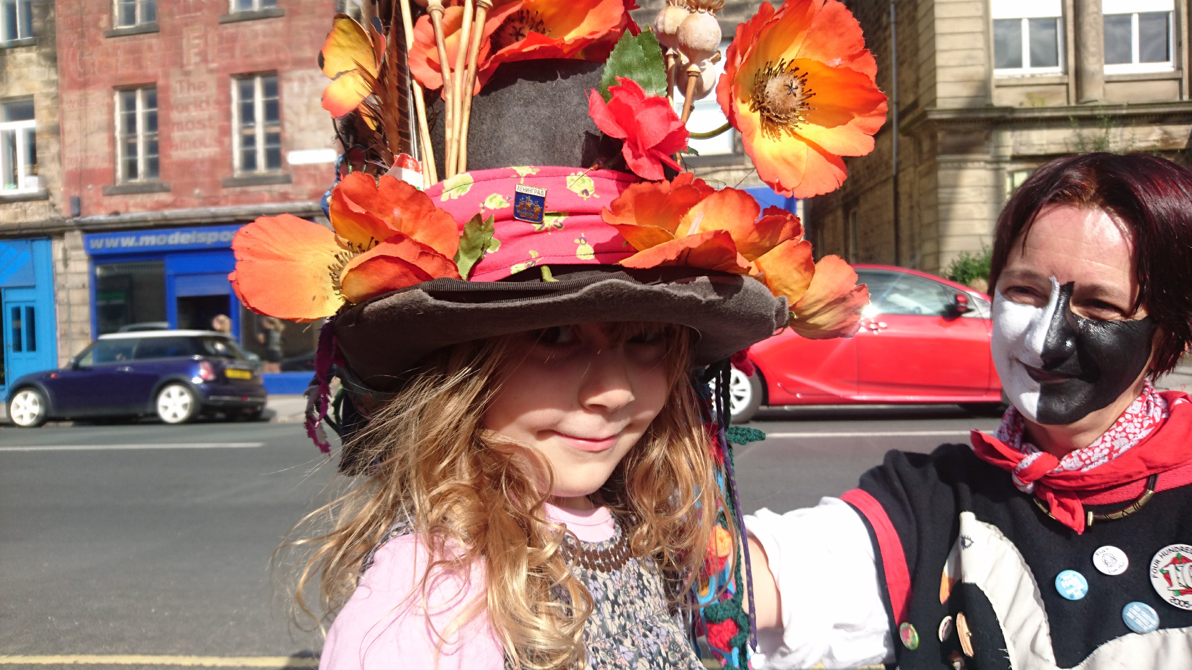 A child in a large hat decorated with orange flowers stands beside a person in black and white face paint
